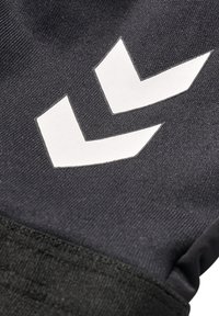 Logo double chevron blanc sur tissu noir texturé, probablement une partie d'une manche ou d'un vêtement de sport.