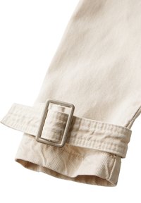 Beige-kankainen hihna, jossa on suorakaiteen muotoinen hopeinen solki. Hihnassa on teksturoitu pinta ja se on valmistettu kestävästä materiaalista.