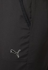 Černé sportovní kalhoty s hladkou texturou, s malým bílým logem Puma na spodní straně a boční kapsou s tkaným detailem.