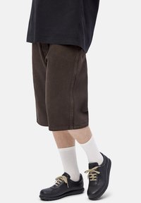 Person wearing dark brown knee-length shorts, white ribbed socks, black leather sneakers with beige laces, and a loose black shirt.