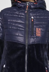 Tummansininen tikattu ja fleecetakki huppulla, edessä vetoketju, vetoketjullinen rintatasku ja oranssi Superdry-logo rinnassa.