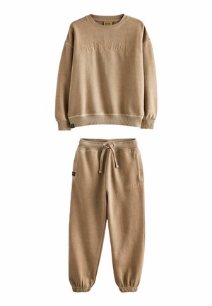 Beige sportovní vršek a odpovídající tepláky. Vršek má kulatý výstřih, žebrované manžety a embosované "SUPERDRY." Tepláky mají šňůrku v pase a zúžené nohavice.