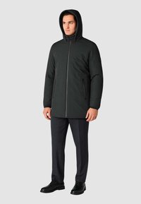 Giacca nera imbottita con cappuccio, zip frontale e due tasche laterali. Indossata sopra pantaloni grigi scuri e scarpe nere. Texture liscia.