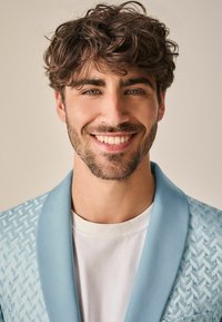 Blazer azzurro chiaro con motivo testurizzato, con risvolti, indossato sopra una t-shirt bianca. Il soggetto ha capelli ricci castani e una barba.