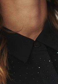Black shirt with a collar, featuring textured fabric and a black button. The shirt is adorned with subtle, small shiny dots.