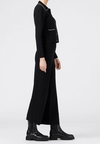 Černý oblek na míru se štíhlým sakem s bílým lemováním a dlouhými širokými kalhotami. Doplněno o elegantní černé kotníkové boty.