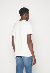 Balta kokvilnas T-kreklu ar apaļu kakla izgriezumu un īsām piedurknēm, ar vienkāršu dizainu. Uzvilkta kopā ar gaiši zilām džinsu biksēm, skatīta no aizmugures.