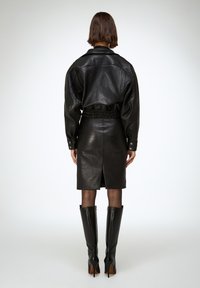 Mujer de espaldas a la cámara, vestida con chaqueta de cuero negra, falda a la altura de las rodillas, medias transparentes y botas negras de tacón alto hasta la rodilla.