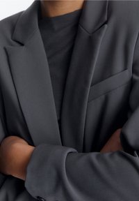 Blazer gris foncé à coupe ajustée, avec des revers crantés, une poche poitrine et des poignets à boutons. Texture de tissu lisse.