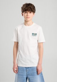 Valkoinen puuvillapaita, jossa on vihreä painatus, jossa lukee "Hyvä Elämä" ja kukka-aiheisia koristuksia vasemmalla rinnalla. Lyhyet hihat, pyöreä kaula-aukko.