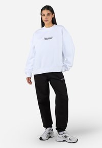 Witte sweatshirt met een ronde hals en geborduurde tekst, gecombineerd met zwarte sweatpants en grijze sneakers. Ontspannen pasvorm, casual stijl.