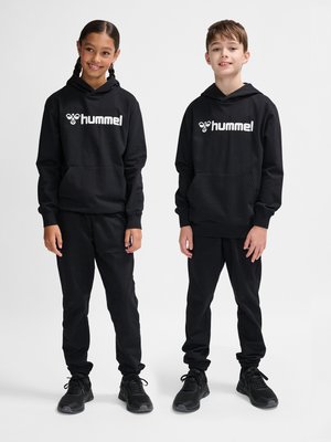 Deux enfants debout côte à côte portant des sweats à capuche noirs hummel assortis, des pantalons noirs et des baskets noires sur un fond uni.