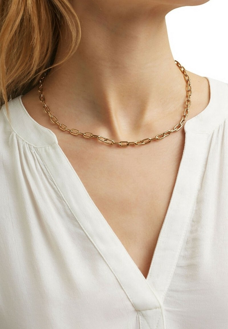 Collier en chaîne d'or avec des maillons allongés porté autour du cou d'une femme, sur un chemisier blanc à col en V.