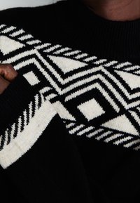 Svart stickad tröja med ett geometriskt vitt mönster och kontrasterande texturer på ärmarna och halsringningen.