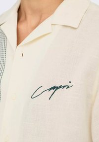 Camisa de manga corta color crema, hecha de una tela ligera, con un acabado texturizado y un logo bordado en verde en el pecho.