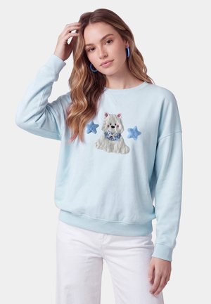 Giovane donna con capelli mossi che indossa una felpa azzurro chiaro con un design di cane e stelle, abbinata a pantaloni bianchi e orecchini a cerchio blu.