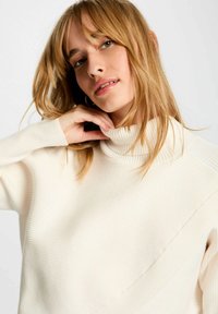 Maglione a collo alto crema con texture a coste, caratterizzato da un elemento di design diagonale e maniche lunghe. Tessuto morbido e vestibilità comoda.