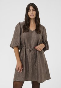 Frau mit langen dunklen Haaren, die ein braunes, strukturiertes Kleid mit gepufften Ärmeln und einem schmalen Bindegürtel trägt, steht vor einem schlichten weißen Hintergrund.