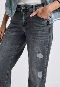 Jean en denim gris foncé avec une coupe décontractée, présentant des déchirures au-dessus du genou droit et une fermeture à bouton classique à la taille.