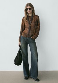 Brun mønstret bluse med lange ærmer og bindebånd ved halsen, kombineret med vidde bukser i mørkeblå og en sort skuldertaske.