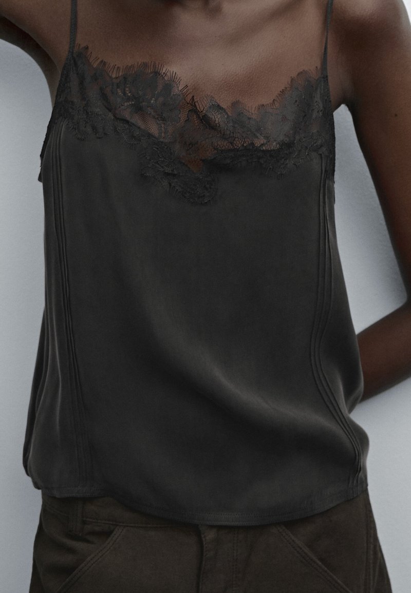 Femme portant un caraco en satin noir avec bordure en dentelle, assorti à un pantalon sombre, vue de la poitrine à la taille.