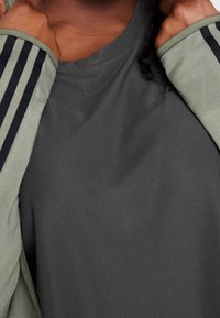 Haut de sport gris en tissu texturé, avec des rayures diagonales noires le long des manches et une coupe ample et décontractée.