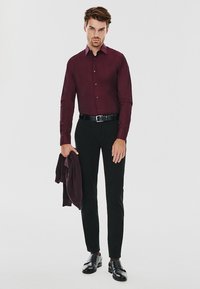 Burgundowa koszula z guzikami, dopasowane czarne spodnie, czarne buty oraz pasujący sweter zarzucony na jedno ramię. Gładki materiał, dopasowany fason.
