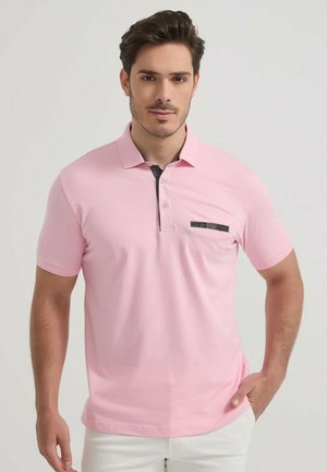 Homme portant un polo rose clair à manches courtes avec un col et une poche bordés de noir, debout devant un fond clair uni.