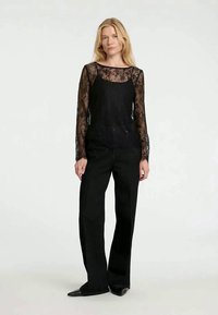 Woman with blonde hair wearing black lace long-sleeve top layered under a black camisole, black wide-leg pants, and black pointed shoes.