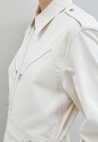 Světle béžový overal s límečkem, dvěma zipovými kapsami na hrudi a kontrastním červeným prošitím. Vyroben z texturované látky.