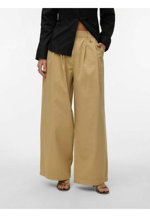 Femme portant un pantalon beige à jambes larges, un haut noir à manches longues, et des sandales à brides, debout avec une main dans la poche.