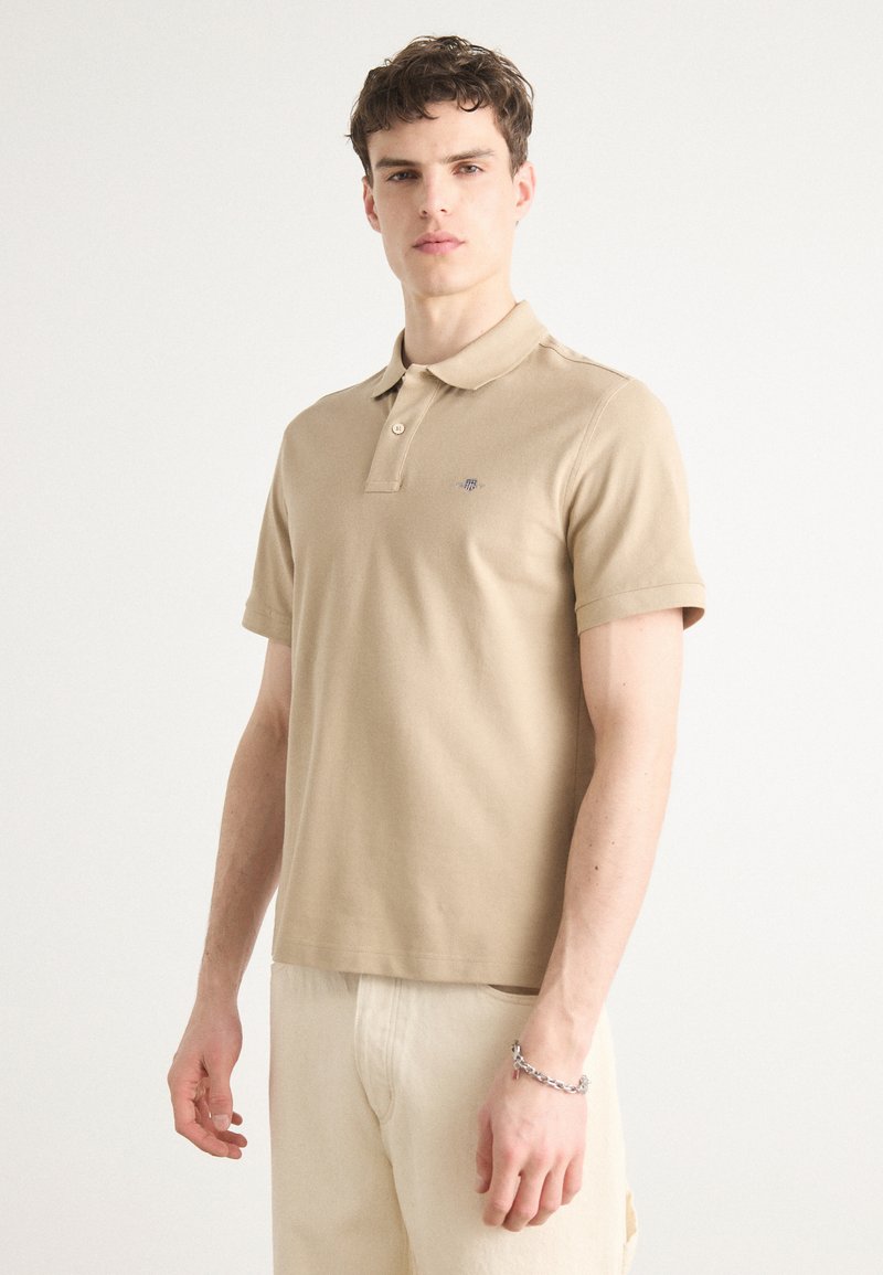 Polo en coton beige à manches courtes, avec un plastron à deux boutons et un petit logo brodé sur la poitrine. Porté avec un pantalon de couleur claire.