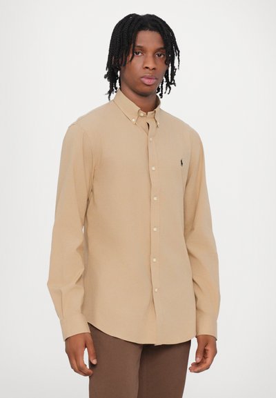 Camisa de botão de manga longa bege com colarinho pontiagudo, feita de um tecido suave, apresentando um pequeno logótipo no lado esquerdo do peito.