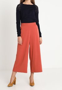 Haut à manches longues bleu marine avec des accents en dentelle et un pantalon large corail. Le modèle porte des chaussures à talons bloc moutarde. Design simple et ajusté.