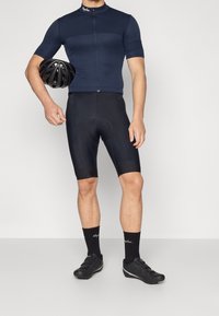 Conjunto de ciclismo: camiseta azul marino de manga corta, pantalones cortos acolchados negros, zapatillas de ciclismo negras y un casco negro sujetado bajo un brazo.