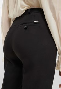Pantaloni neri con una texture liscia, dotati di una tasca posteriore con bottone e un piccolo logo metallico.