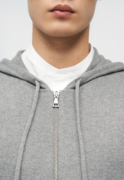 Κοντινή λήψη ενός ατόμου που φοράει λευκό πουκάμισο με γιακά κάτω από γκρι πλεκτό φούτερ με φερμουάρ και κορδόνια, με ορατά τα χείλη και τον λαιμό.