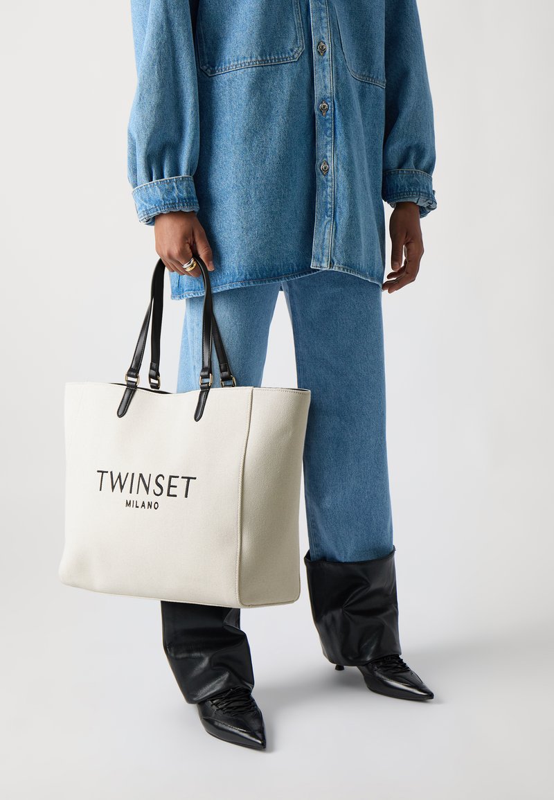Krém színű vászontáska fekete fogantyúkkal és a "TWINSET MILANO" emblémával. Farmer outfittel és fekete, hegyes orrú csizmával párosítva.