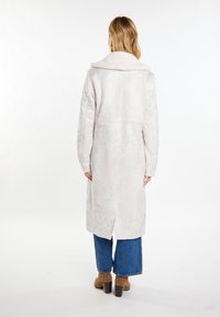 Un lungo cappotto grigio chiaro presenta una texture morbida, un grande colletto e spacchi laterali, abbinato a jeans blu a gamba larga e stivali con tacco marroni.