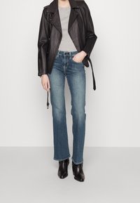 Person som bär en svart läderjacka, grå skjorta, blå jeans och svarta ankla stövlar, står mot en enfärgad bakgrund med ena handen i fickan.