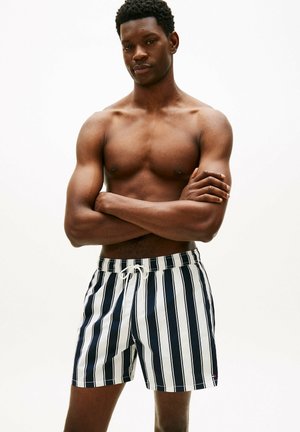 Muskuløs mand stående med krydsede arme iført sort-hvide, lodrette stribede badeshorts mod en ensfarvet hvid baggrund.