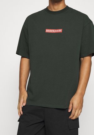 Green cotton t-shirt with a round neck and short sleeves, featuring a rectangular red logo on the chest. Simple, relaxed fit.