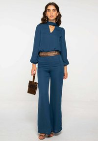 Combinaison turquoise avec un col en V et des manches froncées, dotée d'une large ceinture et de jambes évasées. Pochette ornée d'accents dorés et sandales simples.