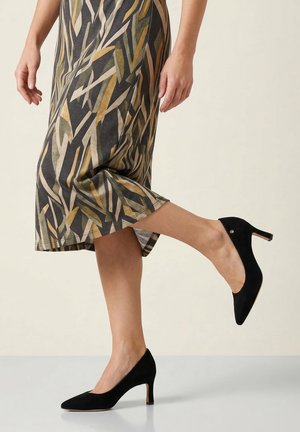 Kobieta w spódnicy midi w wzory i czarnych butach na średnim obcasie, lekko podnosi jedną nogę, aby pokazać boczny profil buta.