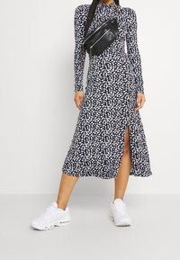 Långärmad svart klänning med vitt prickmönster och sidoklyfta, parat med en svart midjeväska och vita sneakers. Mjuk tygfinish.