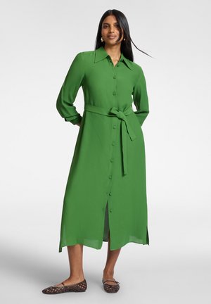 Vrouw met lang zwart haar draagt een groene midi-jurk met knopen, een tailleceintuur en kraag, gecombineerd met bruine gevlochten platte schoenen tegen een effen achtergrond.