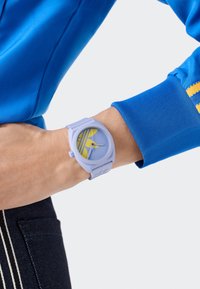 Lilla silikoonkell ümmarguse näo ja sinise kellanäidikuga, millel on kollased triibud ja aktsendid. Kantud heledate teksadega randmel.