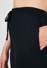 Černé tepláky vyrobené z měkkého materiálu, s páskem na stažení a vzorem diamantů poblíž kapsy. Vhodné pro volnočasové nošení.