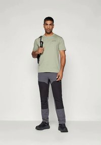 Πράσινο κοντομάνικο πουκάμισο και γκρι-μαύρο παντελόνι εξωτερικού με αντίθετα πάνελ και λογότυπο. Το μοντέλο φοράει μαύρα παπούτσια και κρατάει μια τσάντα.