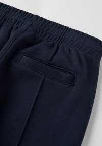 Marineblauwe sportbroek met een elastische tailleband en een achterzak. Geweven stof met een ruitpatroon, ontworpen voor comfort en bewegingsvrijheid.